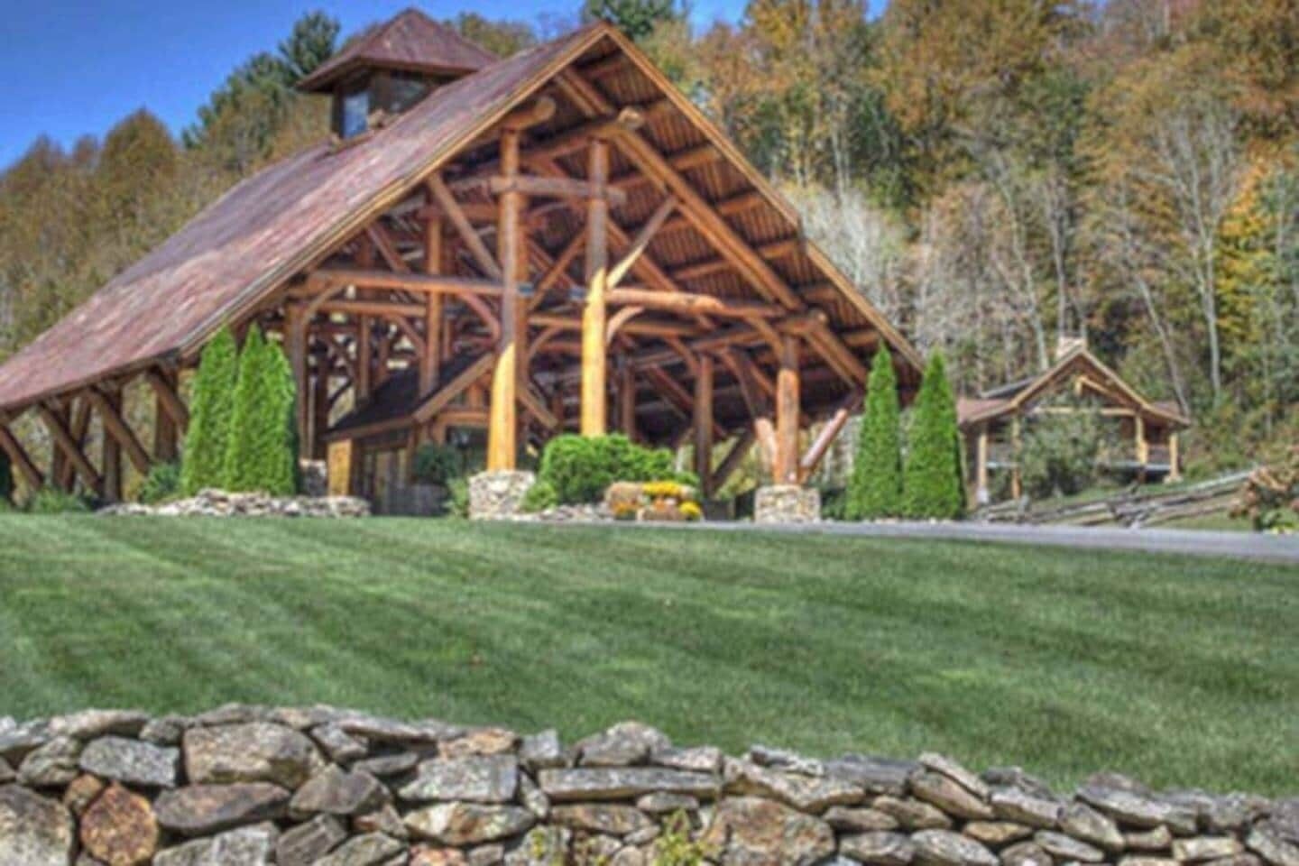 Un bâtiment rustique en bois, de style grange, avec des fondations en pierre, entouré d’un mur de pierre et d’une pelouse bien entretenue.
