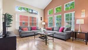 A modern living room with two grey sofas, a coffee table, and a TV stand. There are large windows with multiple panes, allowing natural light to enter. - "The Beauty", a SPLENDID Townhome Near Disney - Free WiFi, sleeps 8 comfortably! (Kissimmee)