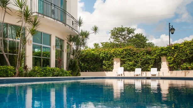 Una piscina con sillas de descanso blancas, rodeada de vegetación exuberante y un edificio con ventanas amplias y un balcón.