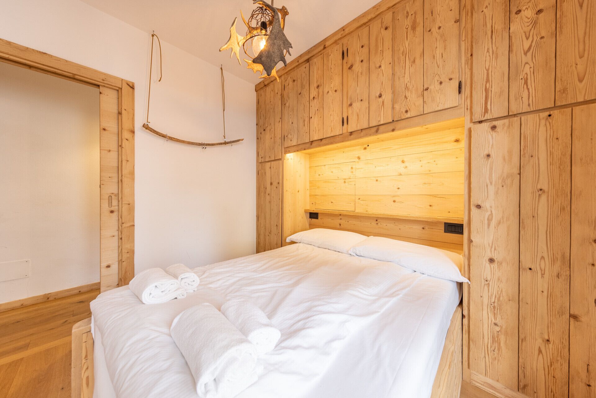 A wooden-paneled room with a bed, a wall-mounted light fixture, and a wooden door.