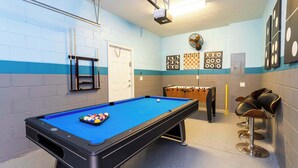 Uma mesa de bilhar com bolas, uma mesa de futebol de mesa e uma mesa de sinuca em um ambiente com paredes azuis e brancas.