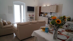 A modern living room with a beige sofa, a dining table with a vase of flowers, and a kitchen area with wooden cabinets and a microwave.