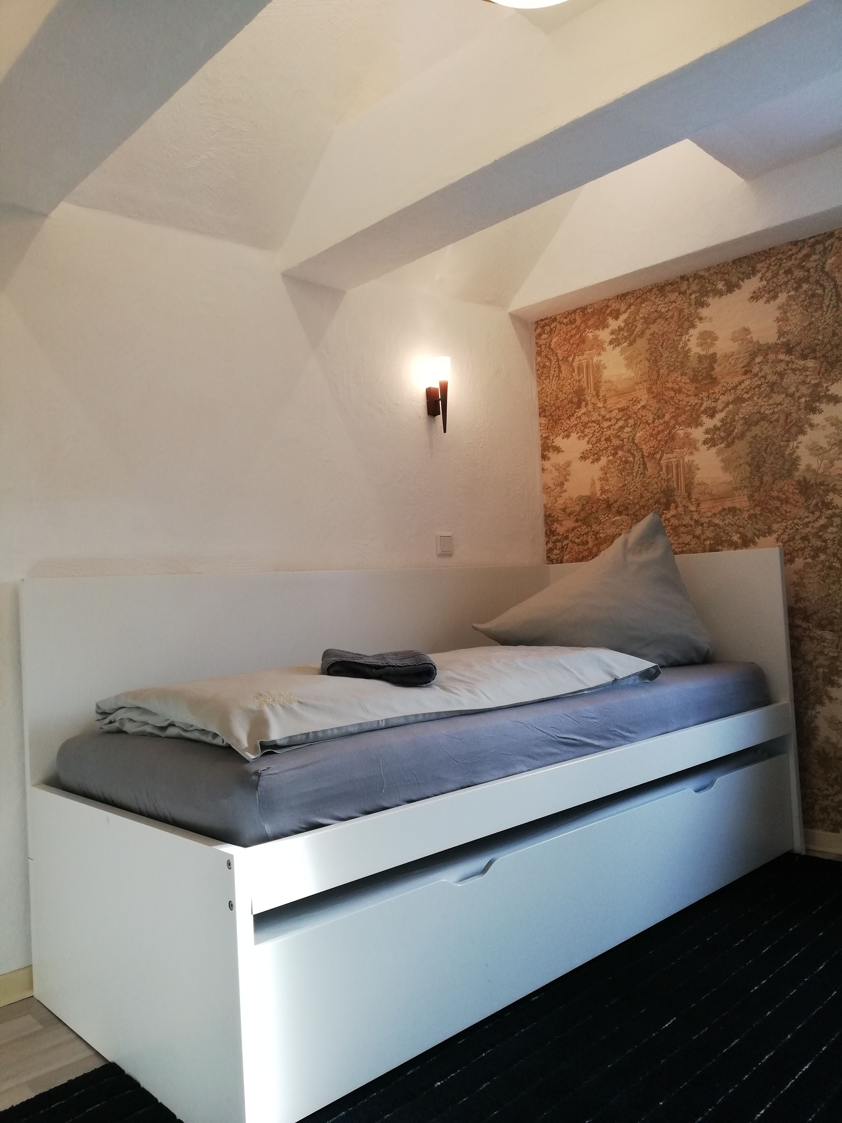 A small, modern bedroom with a white bed, a nightstand, a wall-mounted light, and a patterned wallpaper.