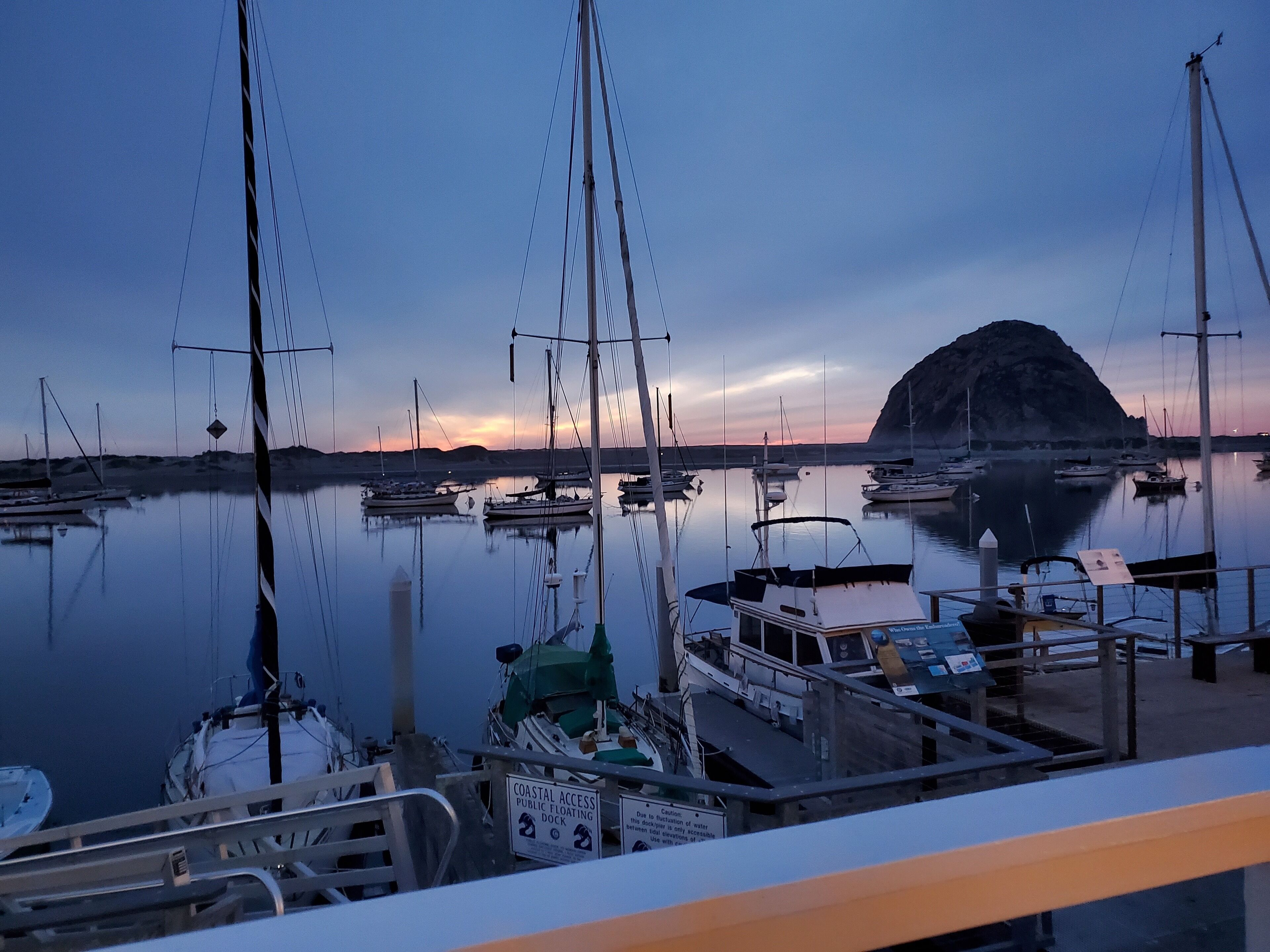 A marina with boats docked, a rocky island in the background, and a sunset.