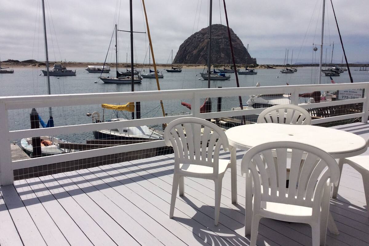 Eine Terrasse mit weißen Kunststoffstühlen und einem Tisch, mit Blick auf einen Yachthafen mit Segelbooten und einer felsigen Insel in der Ferne.