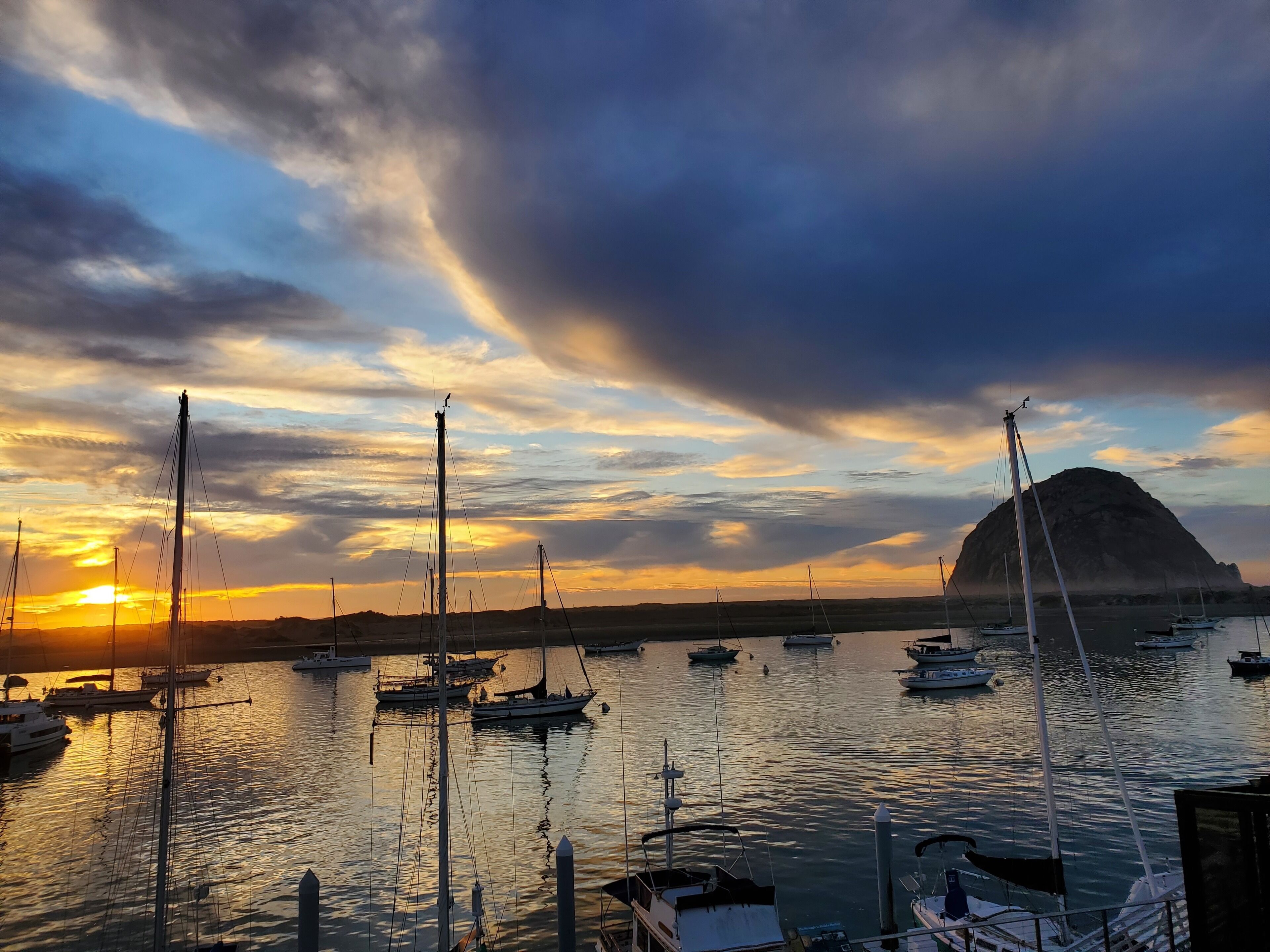 Een jachthaven met meerdere zeilboten afgemeerd, een opvallende berg op de achtergrond en een zonsonderganglucht.