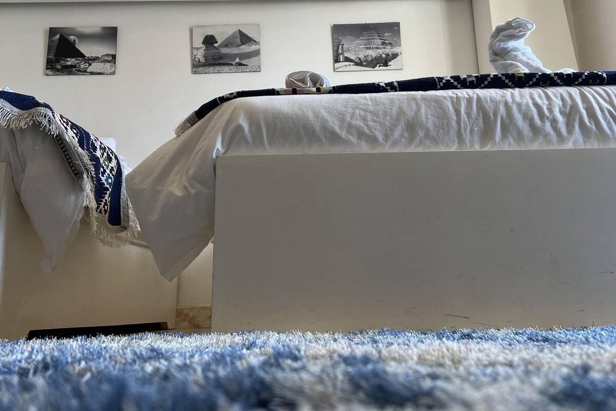 A bedroom with a bed, a white wall with framed pictures, a ceiling light, and an air conditioning unit.