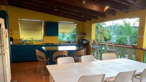 A kitchen with blue cabinets, a white table with chairs, and a balcony with a view of palm trees. - The Waterfront Inn - Hostel (English Harbour)