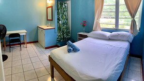 A bedroom with a bed, a desk, a chair, a small cabinet, a window with curtains, and a fan. - The Waterfront Inn - Hostel (English Harbour)