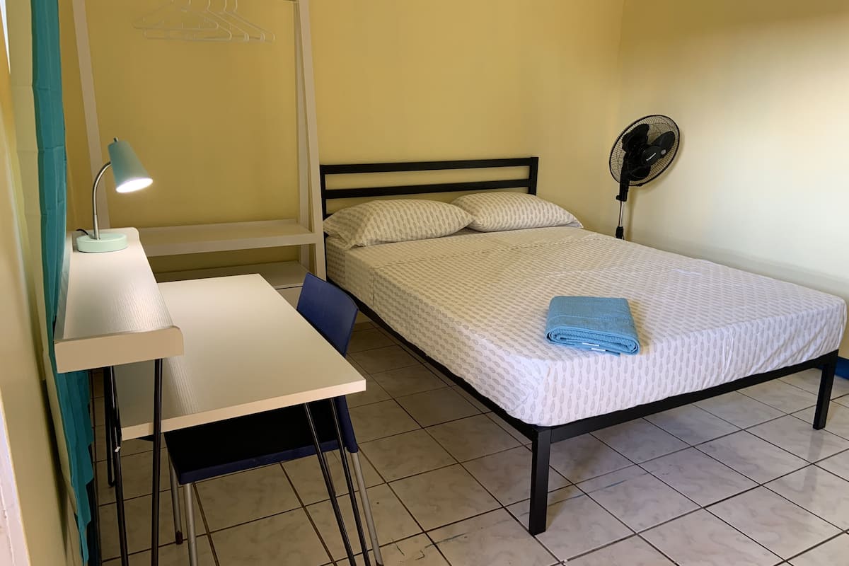 A bedroom with a bed, a desk with a chair, a hanging clothes rack, and a standing fan.