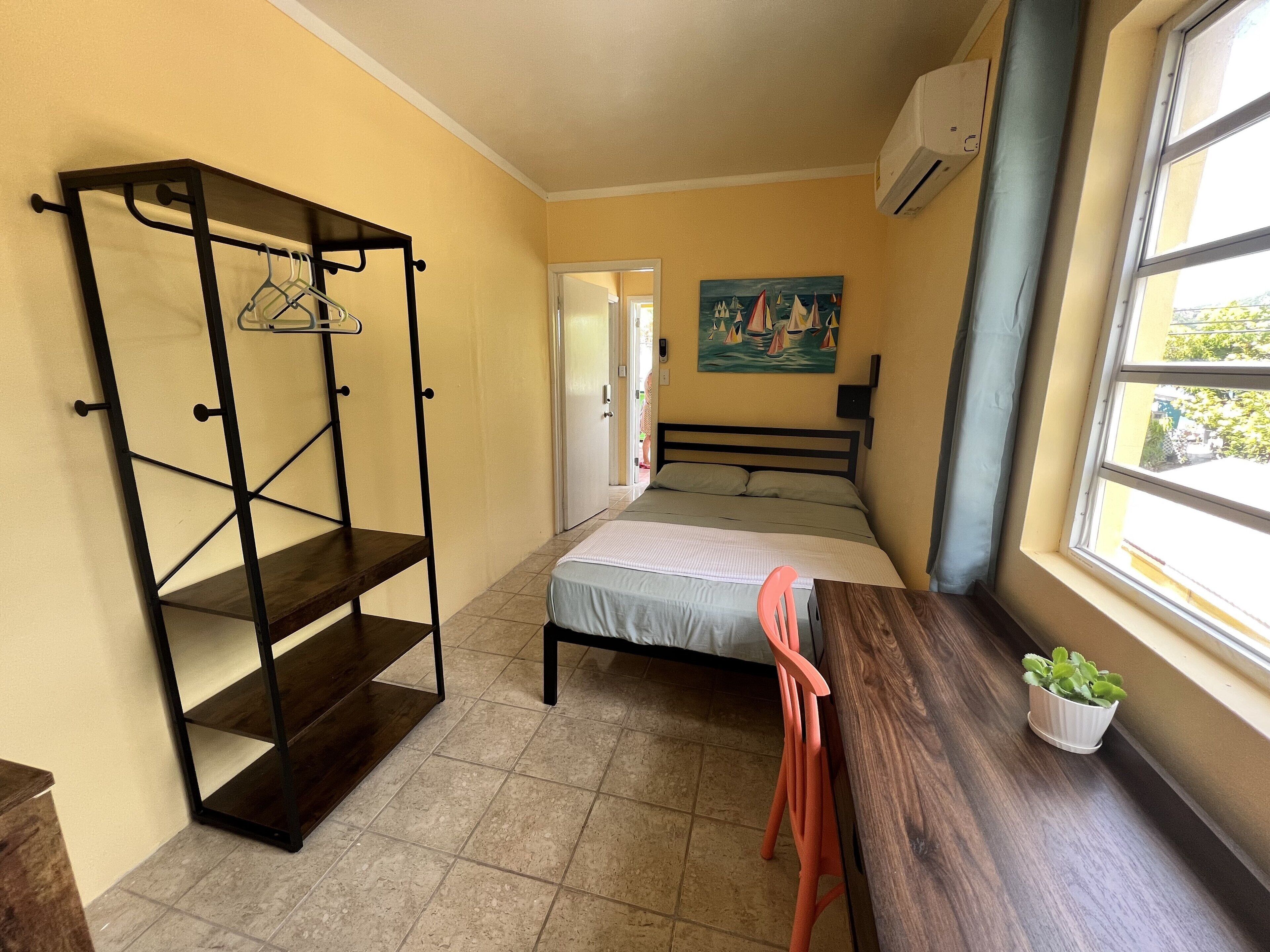 A small, well-lit room with a bed, a chair, a shelf unit, and a window with a view of the outside.