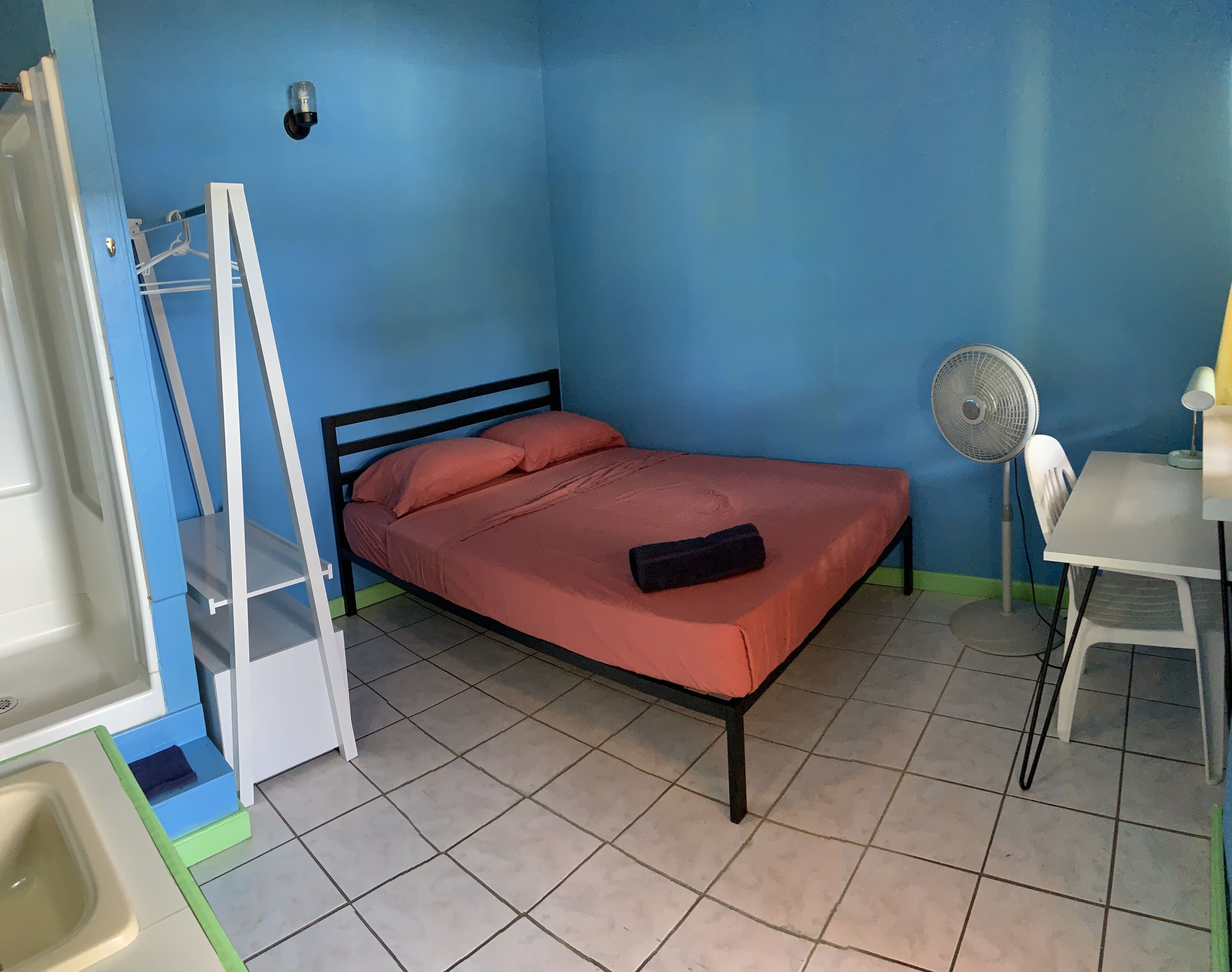 A small, single-bed room with a blue wall, a white desk, a fan, and a small table with a lamp.