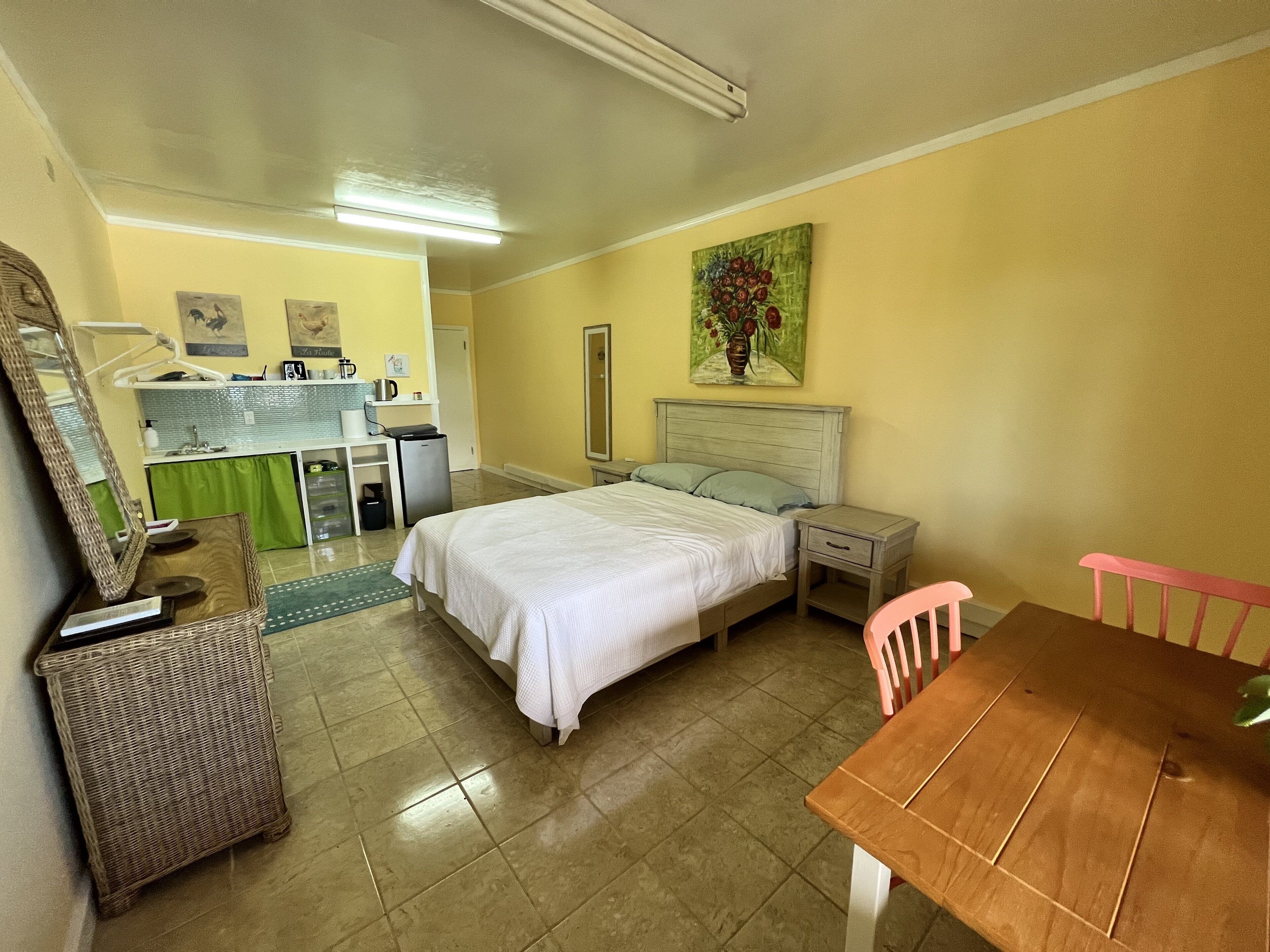A hotel room with a bed, a desk, a chair, a small table, a mirror, a green curtain, and a painting on the wall.