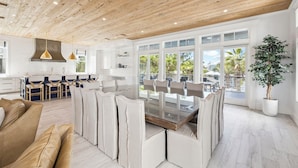 A spacious dining area with a glass-top table and multiple chairs, a wooden ceiling, and large windows with a view of palm trees. - Seas Above the Rest - Short Walk to the Beach! Large Resort Style Private Pool! (Destin)