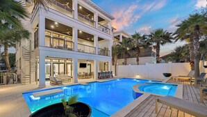 A multi-story building with a swimming pool, outdoor seating, and palm trees. - Seas Above the Rest - Short Walk to the Beach! Large Resort Style Private Pool! (Destin)