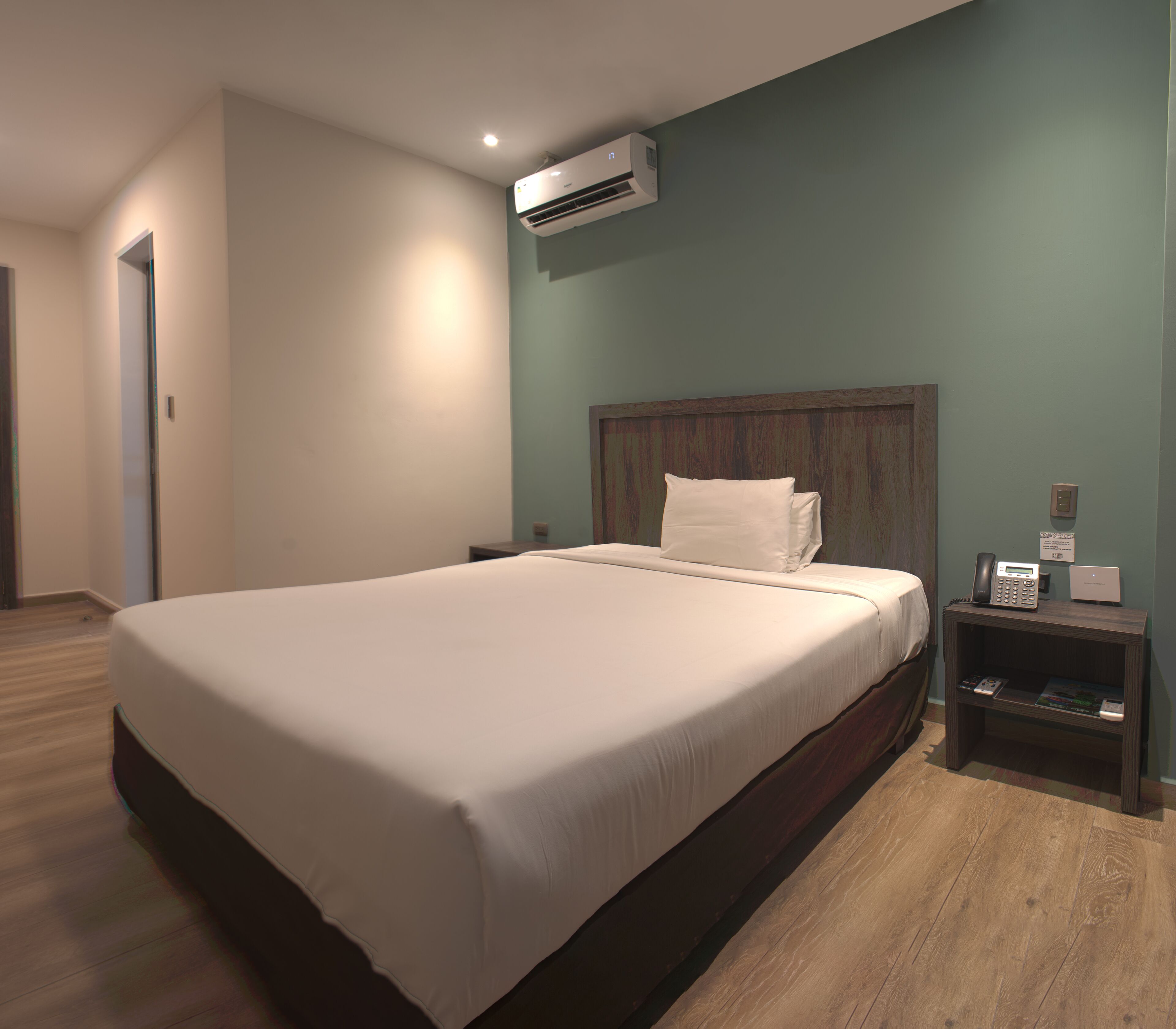 Una habitación de hotel moderna con una cama grande, cabecera de madera, mesita de noche con teléfono y lámpara, y un equipo de aire acondicionado en la pared.