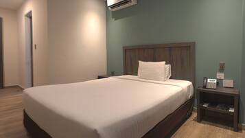 Una habitación de hotel moderna con una cama grande, cabecera de madera, mesita de noche con teléfono y lámpara, y un equipo de aire acondicionado en la pared.