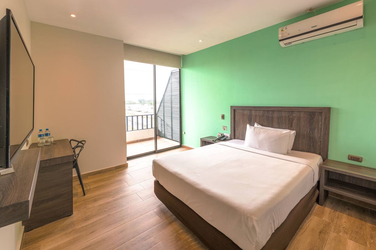 A hotel room with a large bed, a desk with a chair, a nightstand, and an air conditioning unit.