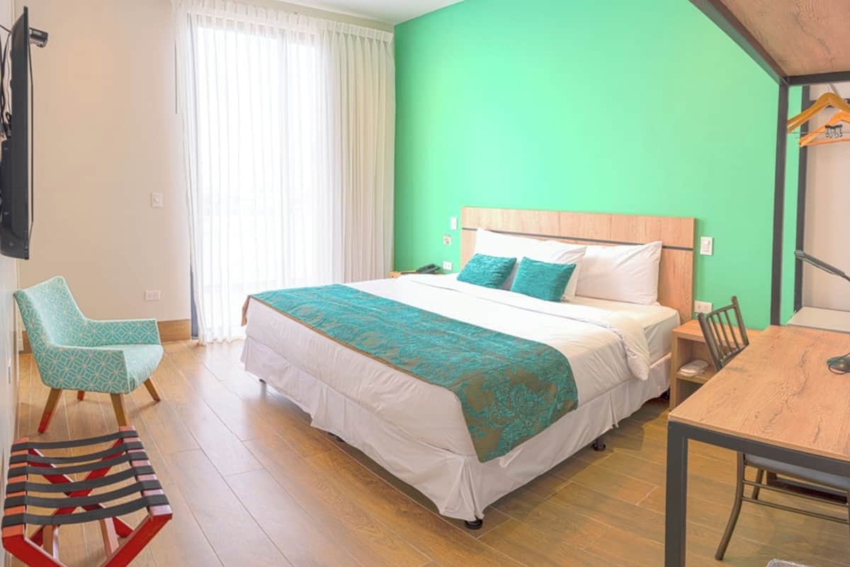 A hotel room with a large bed, a wooden headboard, a green wall, a wooden floor, a chair, a table, and a TV mounted on the wall.