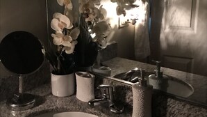 Ein Badezimmer mit einer Granitplatte, einem weißen Waschbecken, einem Spiegel, einer Vase mit weißen Blumen und einem Handtuchspender.