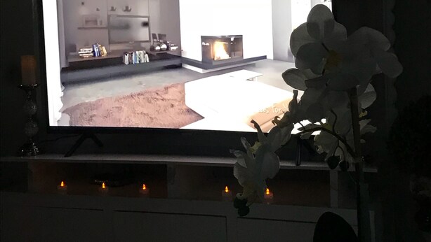 Ein Fernseher zeigt einen Raum mit Kamin, Küchenbereich und einem Esstisch mit Stühlen.