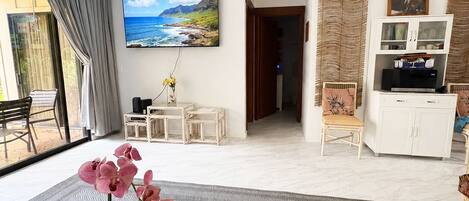 A modern living room with a ceiling fan, a TV mounted on the wall, a dining table with a vase of flowers, and a view of the ocean through a large window.
