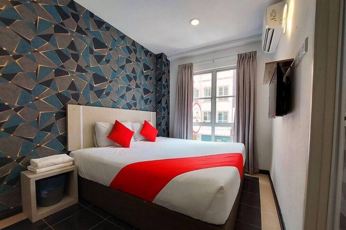 A hotel room with a geometric-patterned wall, a bed with red and white bedding, a bedside table with a towel, and a window with curtains.