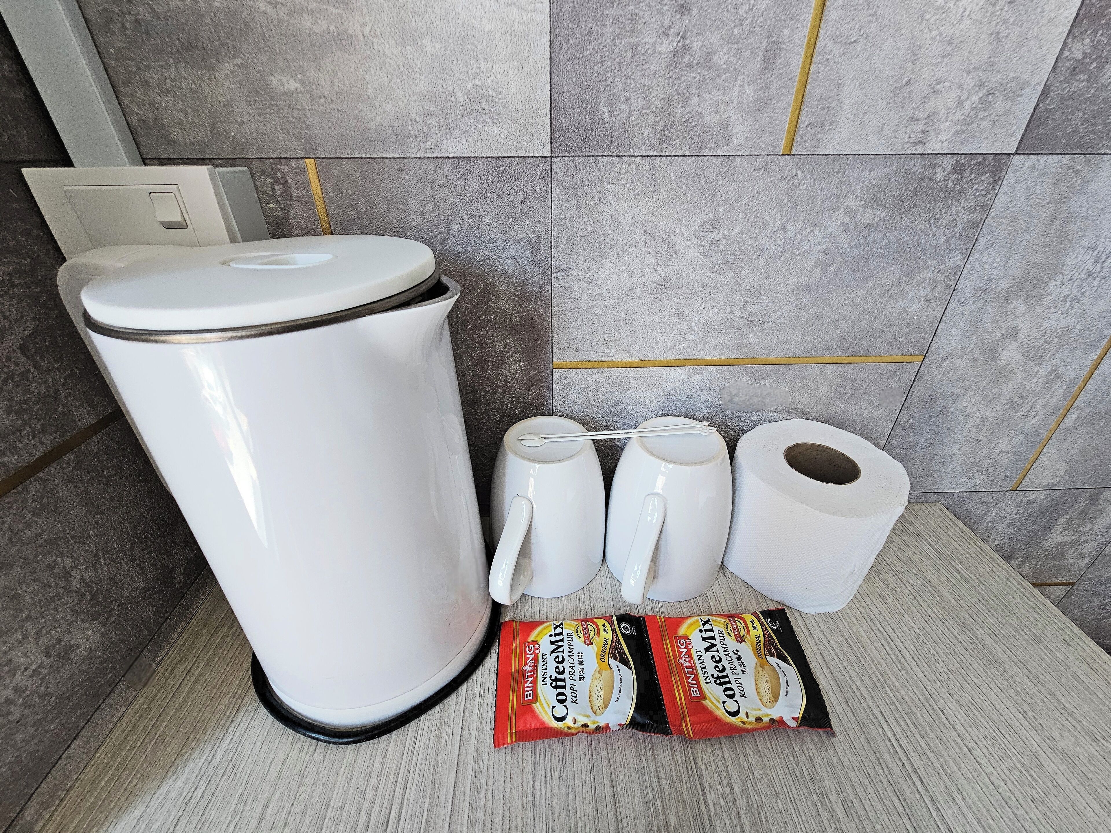 Una cafetera, dos tazas, dos rollos de papel higiénico y dos sobres de café instantáneo sobre una encimera con salpicadero de azulejos.