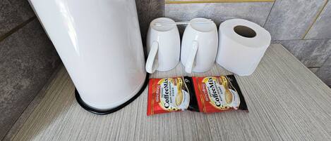 Una cafetera, dos tazas, dos rollos de papel higiénico y dos sobres de café instantáneo sobre una encimera con salpicadero de azulejos.