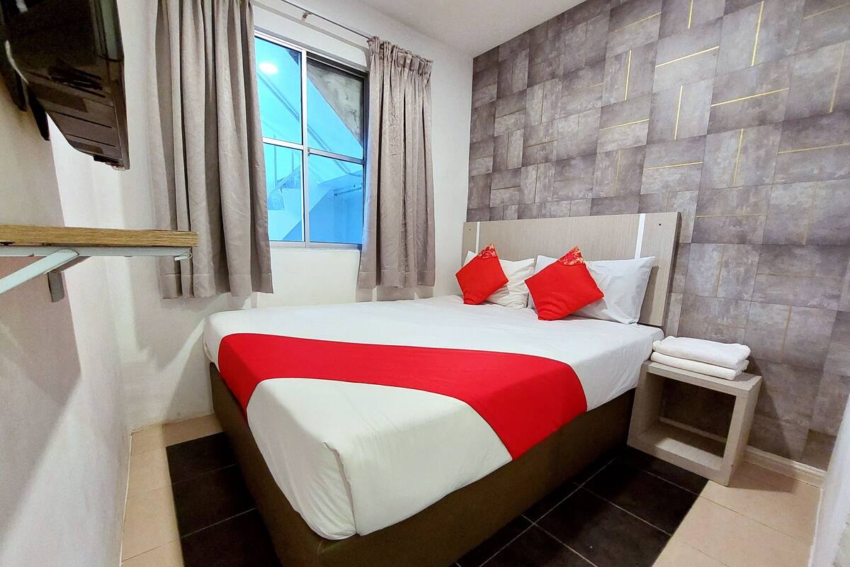 A neatly made bed with a red and white bedspread, a wooden headboard, and two red pillows. There is a window with curtains, a small bedside table with a white pillow, and a tiled floor.