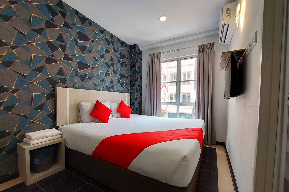 A modern hotel room with a geometric-patterned wall, a large bed with red and white bedding, a bedside table with a towel, and a window with curtains.