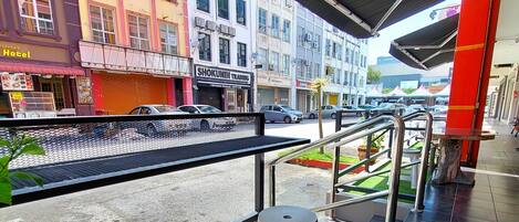 Un espacio de estar al aire libre techado con dos taburetes y una mesa, con vista a una calle con diversas tiendas y edificios.