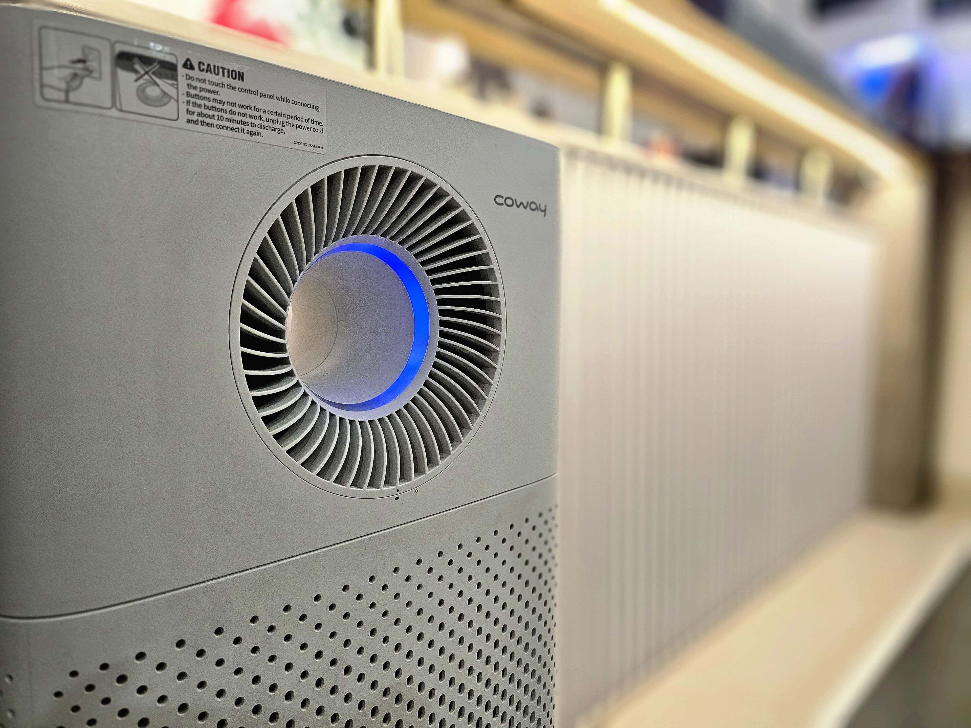 A grey air conditioning unit with a circular vent and a blue light indicator, labeled 'COWAY'.
