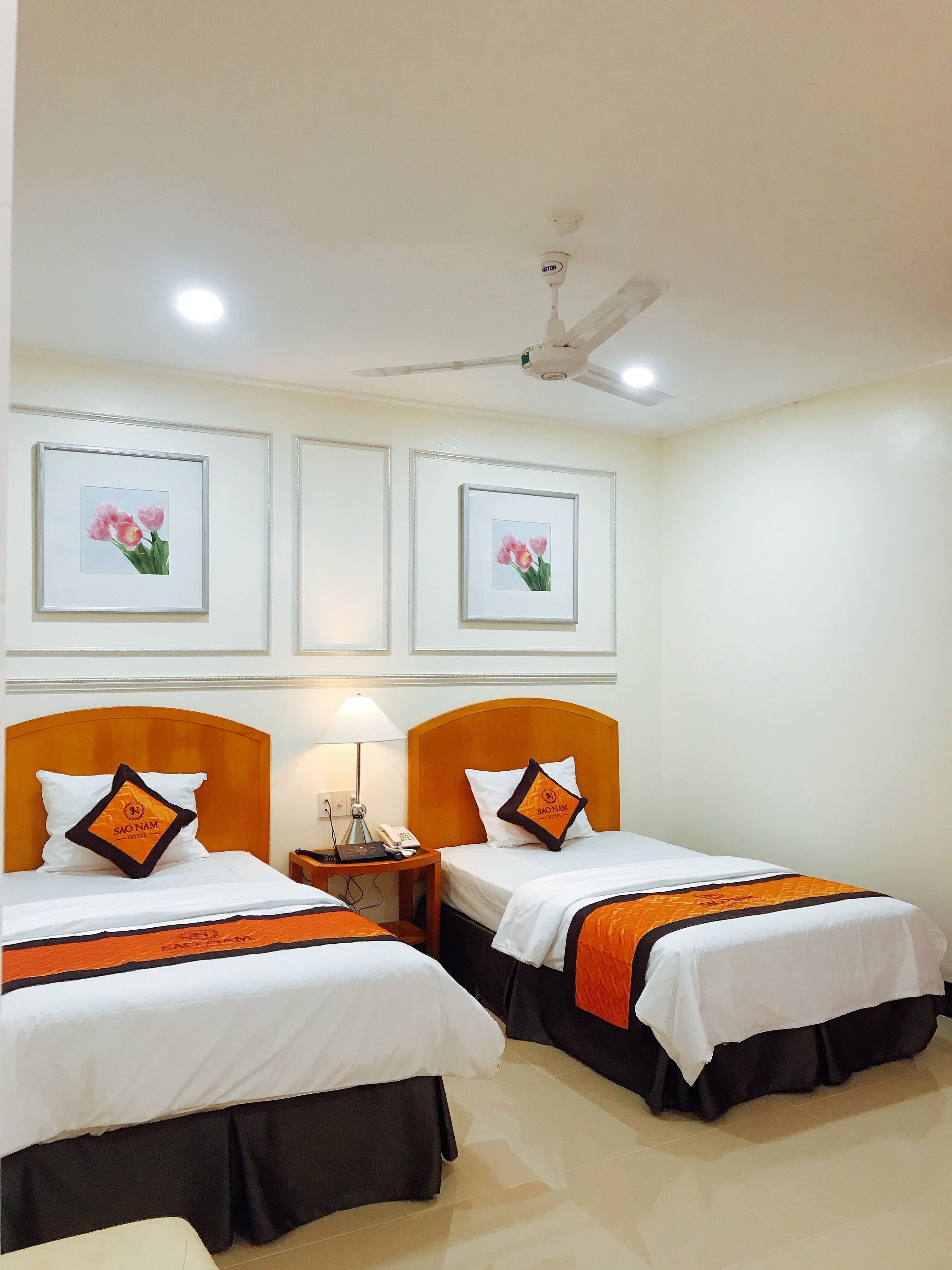 Habitación de hotel con dos camas, cada una con una colcha blanca y naranja, un cabecero de madera y una mesita de noche con lámpara y teléfono. En la pared, encima de las camas, hay cuadros con flores.