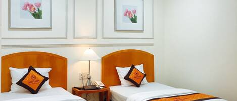 Habitación de hotel con dos camas, cada una con una colcha blanca y naranja, un cabecero de madera y una mesita de noche con lámpara y teléfono. En la pared, encima de las camas, hay cuadros con flores.