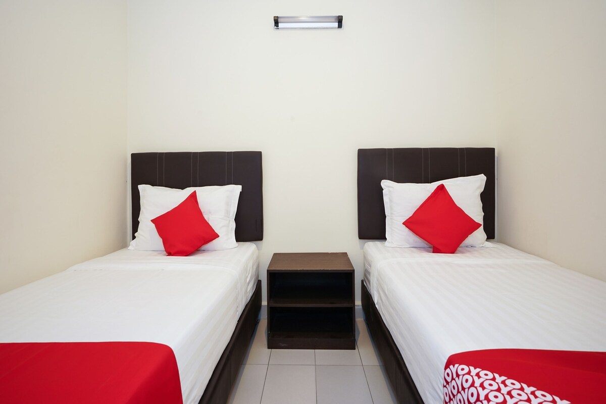 A hotel room with two beds, each with white bedding and red pillows. There is a small wooden nightstand between the beds.