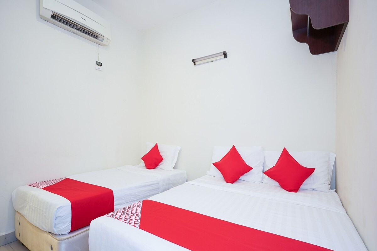 A hotel room with two beds, each with white bedding and red accents, a wall-mounted air conditioner, and a small shelf with a dark brown object.