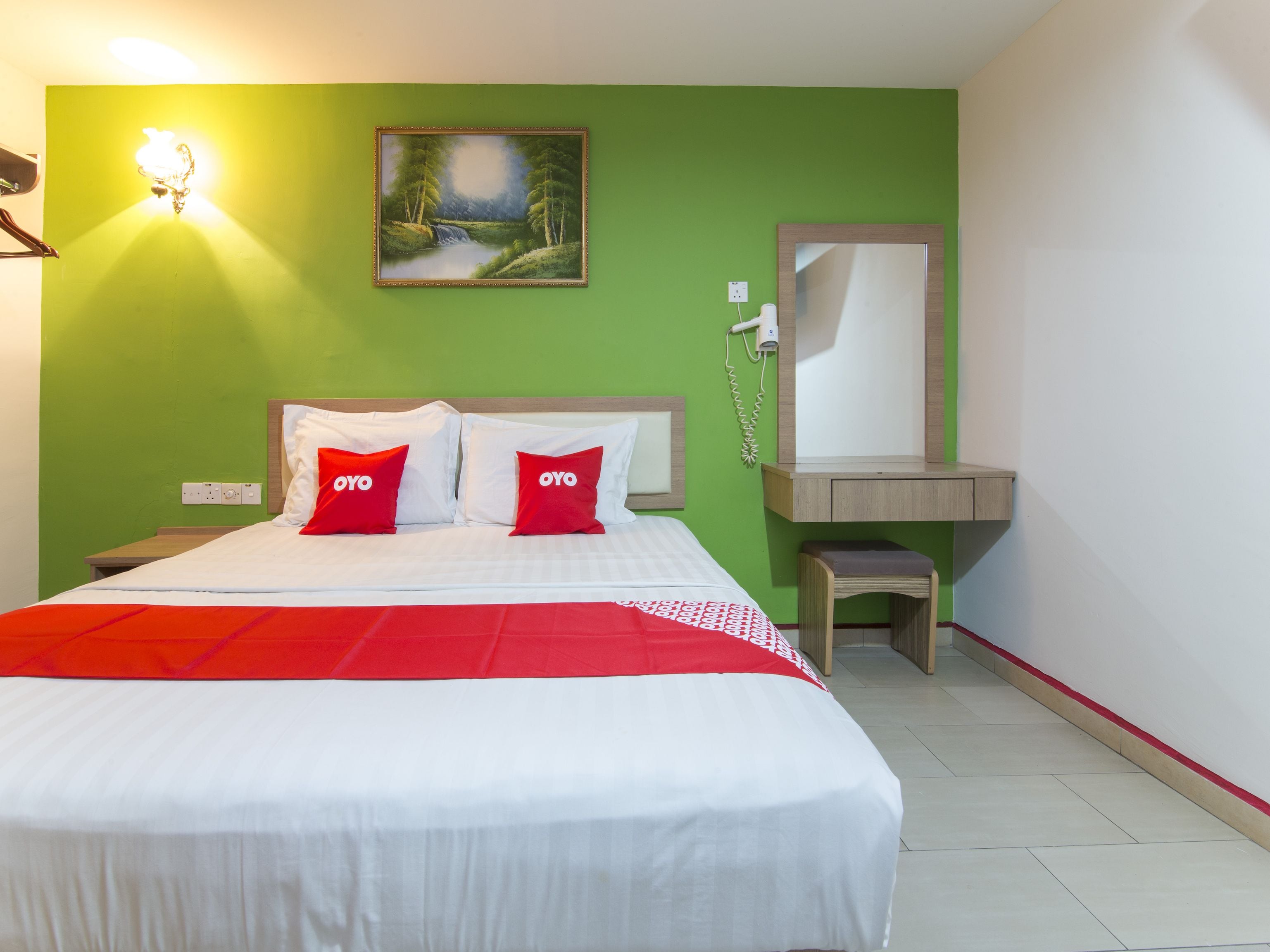 A hotel room with a bed, green walls, a painting of a landscape, a mirror, and a small wooden table.