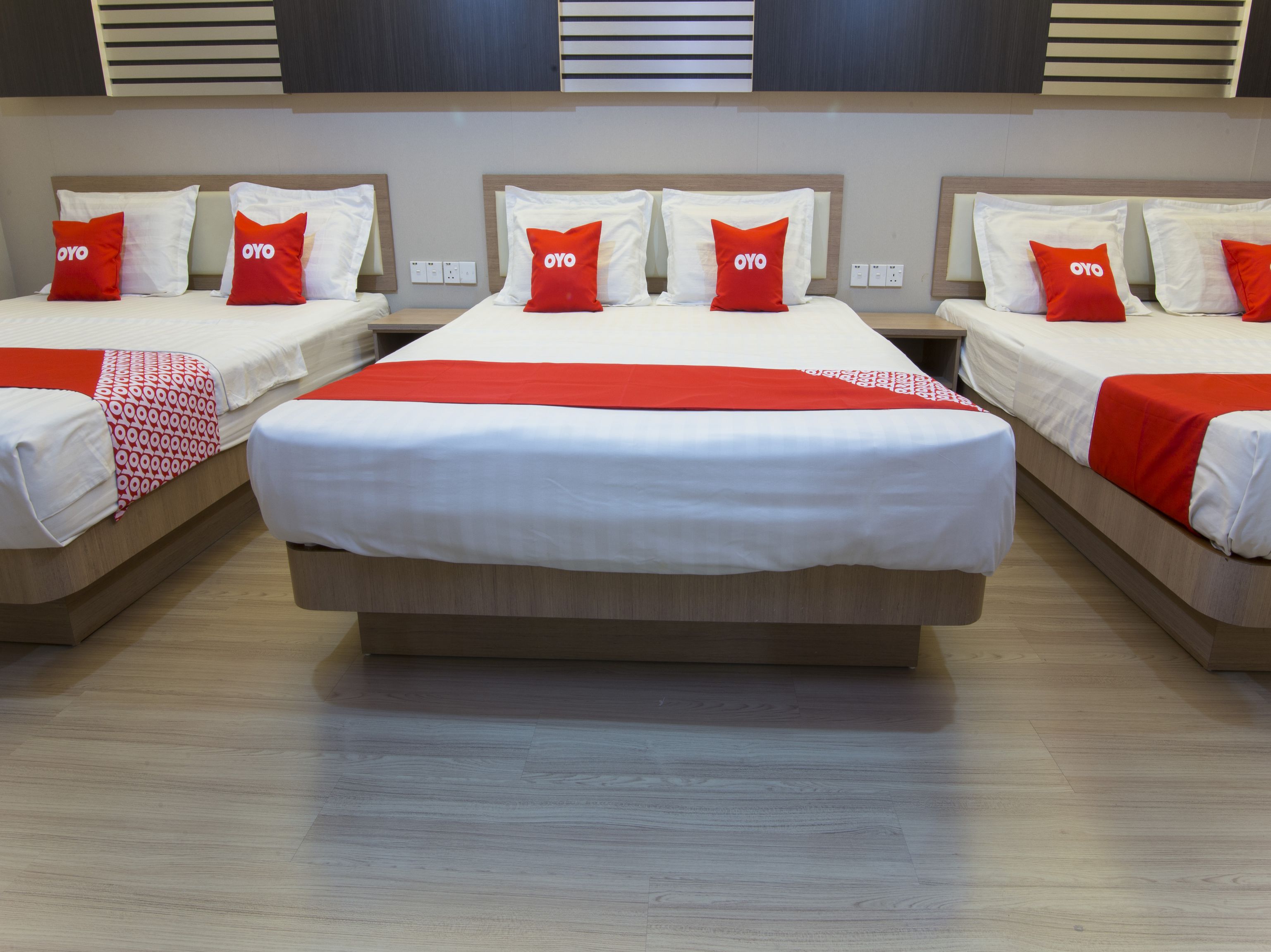 Habitación de hotel con tres camas, cada una con ropa de cama blanca y almohadas rojas, y una manta de colores rojo y blanco.