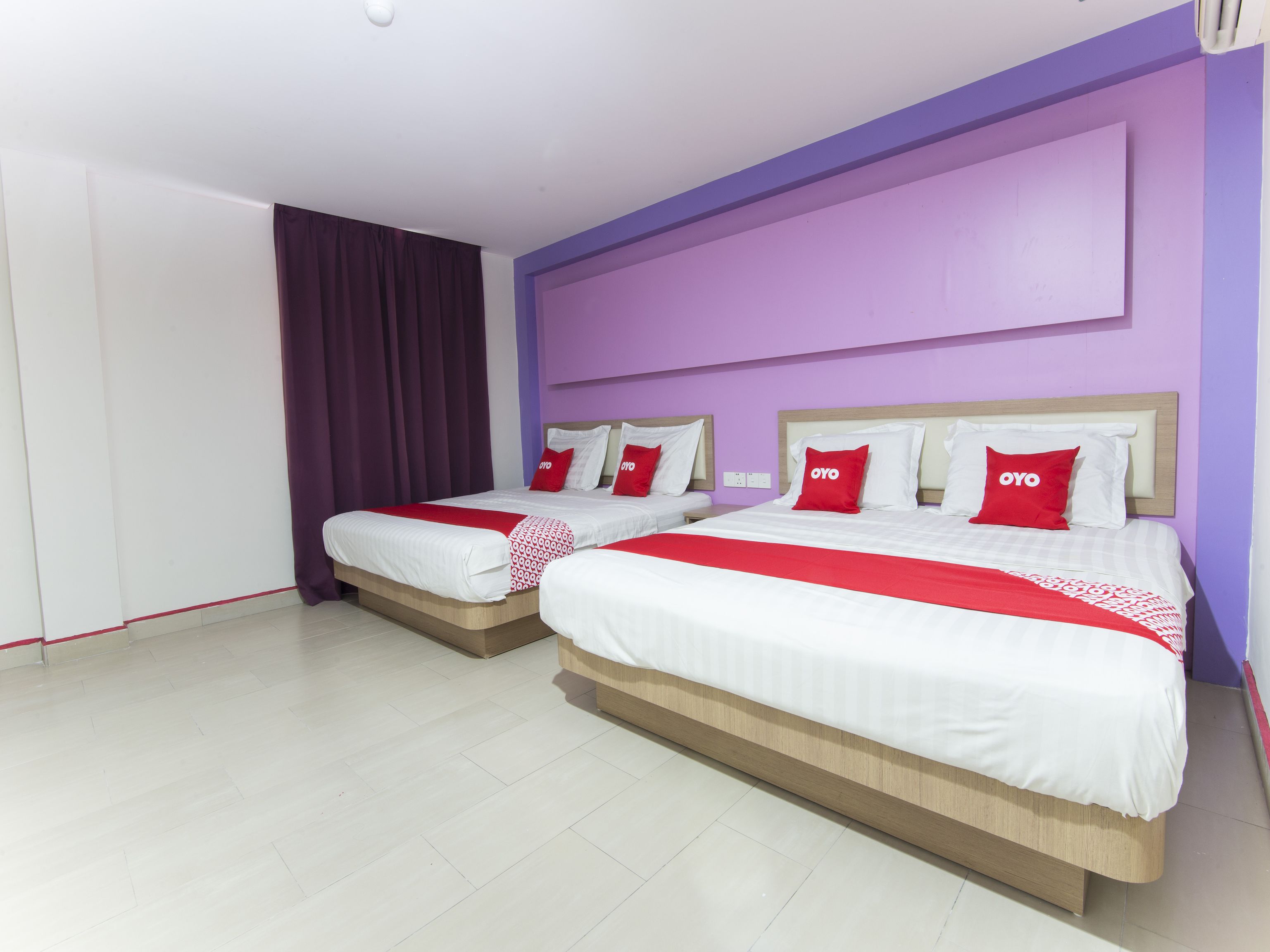 Une chambre d’hôtel avec deux lits, chacun agrémenté de draps blancs et de touches de couleur rouge, un mur d’accent violet et un plancher en bois.