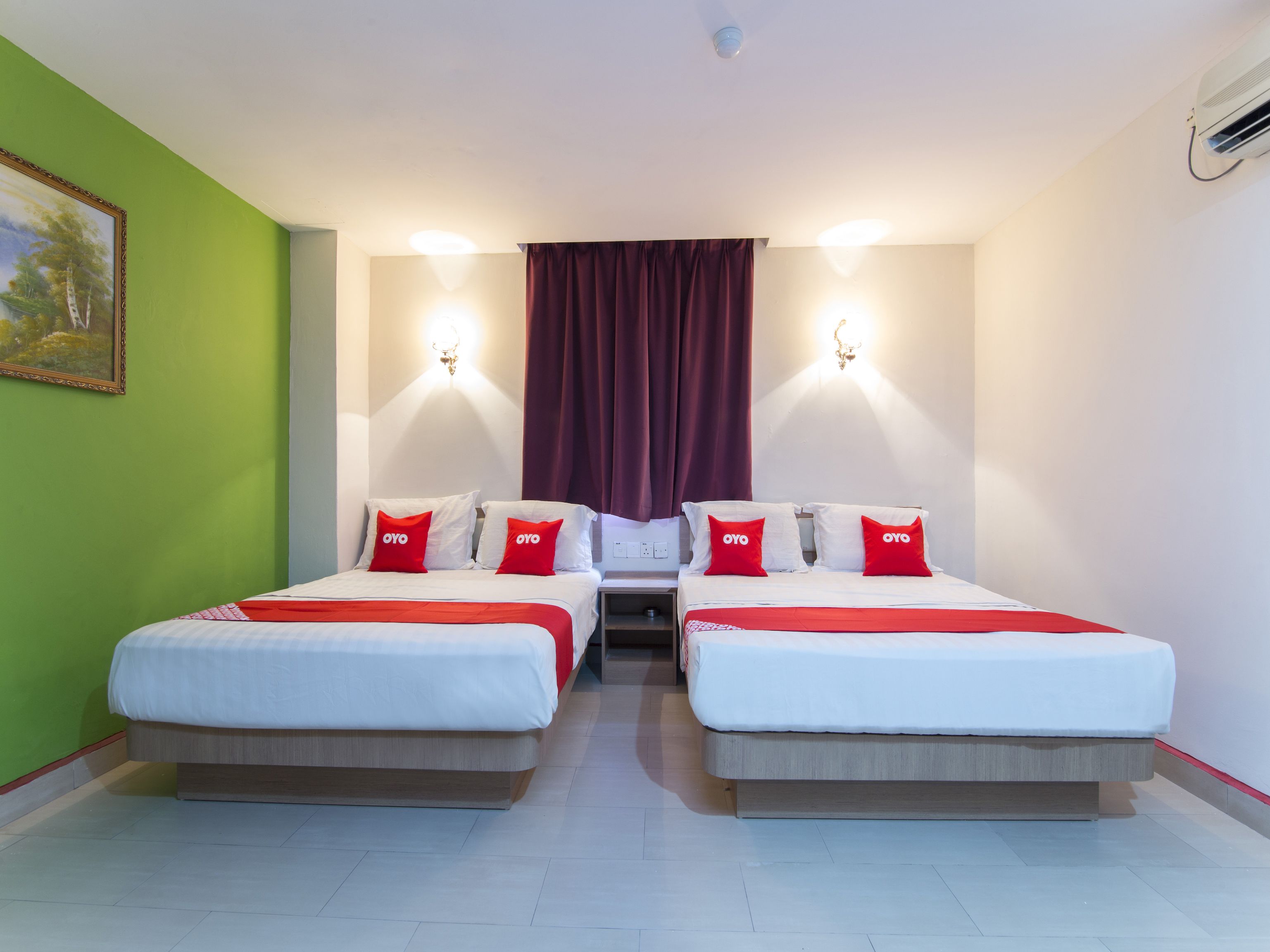 A hotel room with two beds, each with red pillows and a white bedspread. There is a painting on the green wall, a red curtain, and a wall-mounted light fixture.