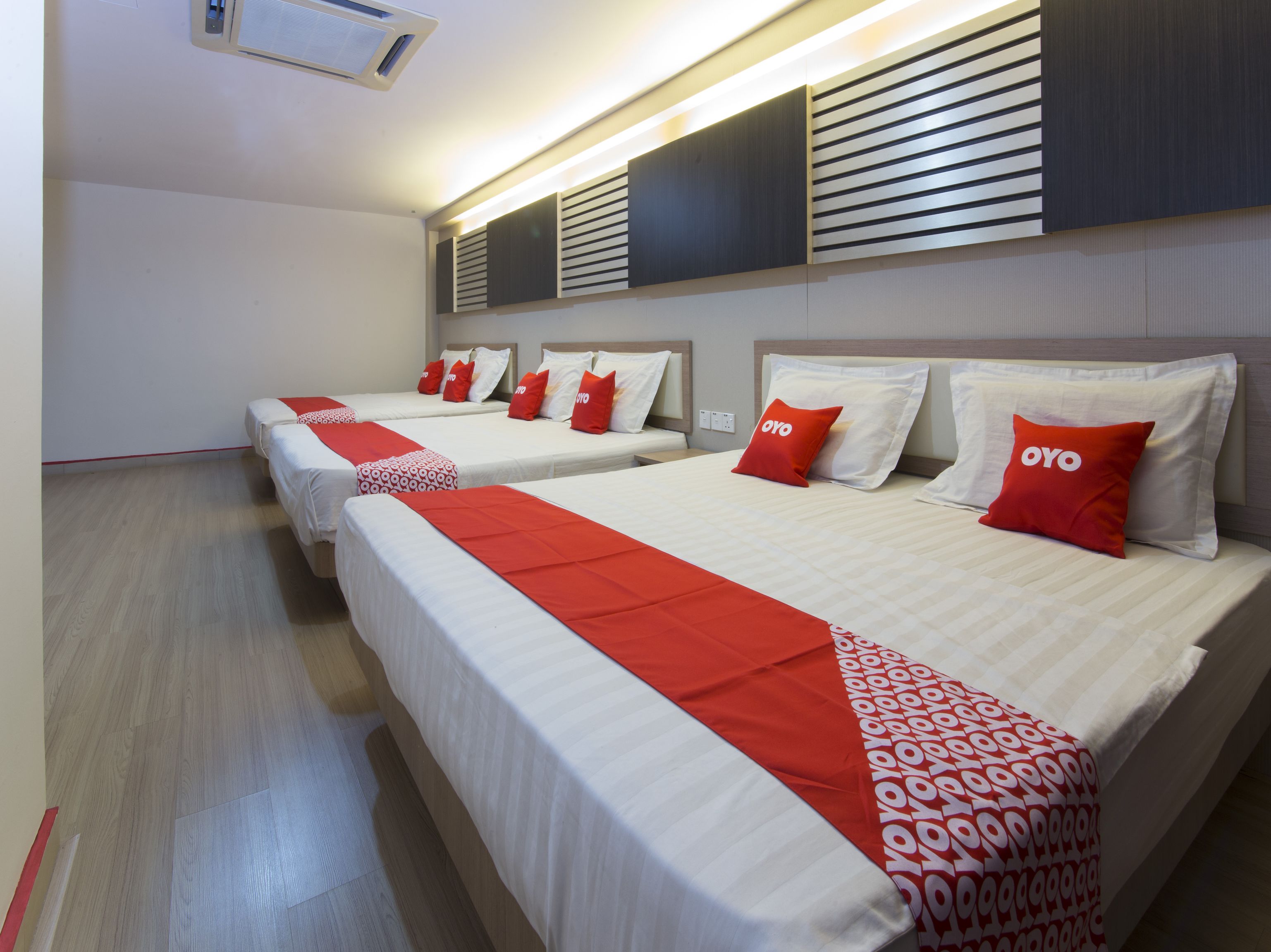 A hotel room with three beds, each with white bedding and red accents, and a modern design with wooden paneling and a built-in air conditioning unit.