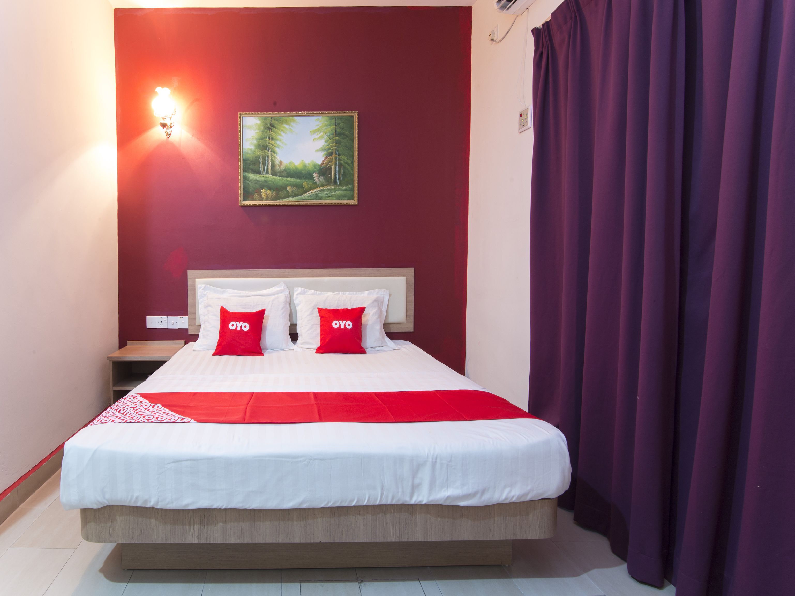 A hotel room with a bed, two red pillows, a painting on the wall, and a purple curtain.