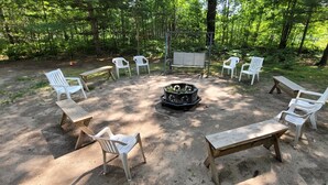 Een ronde zithoek rond een centraal haardvuur in een bebost gebied, met banken en stoelen.