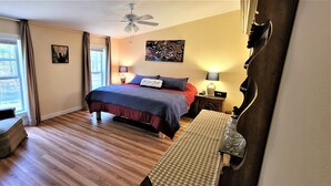 Een slaapkamer met een bed, een stoel, een bureau, een plafondventilator en een raam met gordijnen.