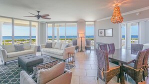 Un salón amplio con un gran ventanal con vistas a la playa, un comedor con sillas de mimbre y un ventilador de techo.