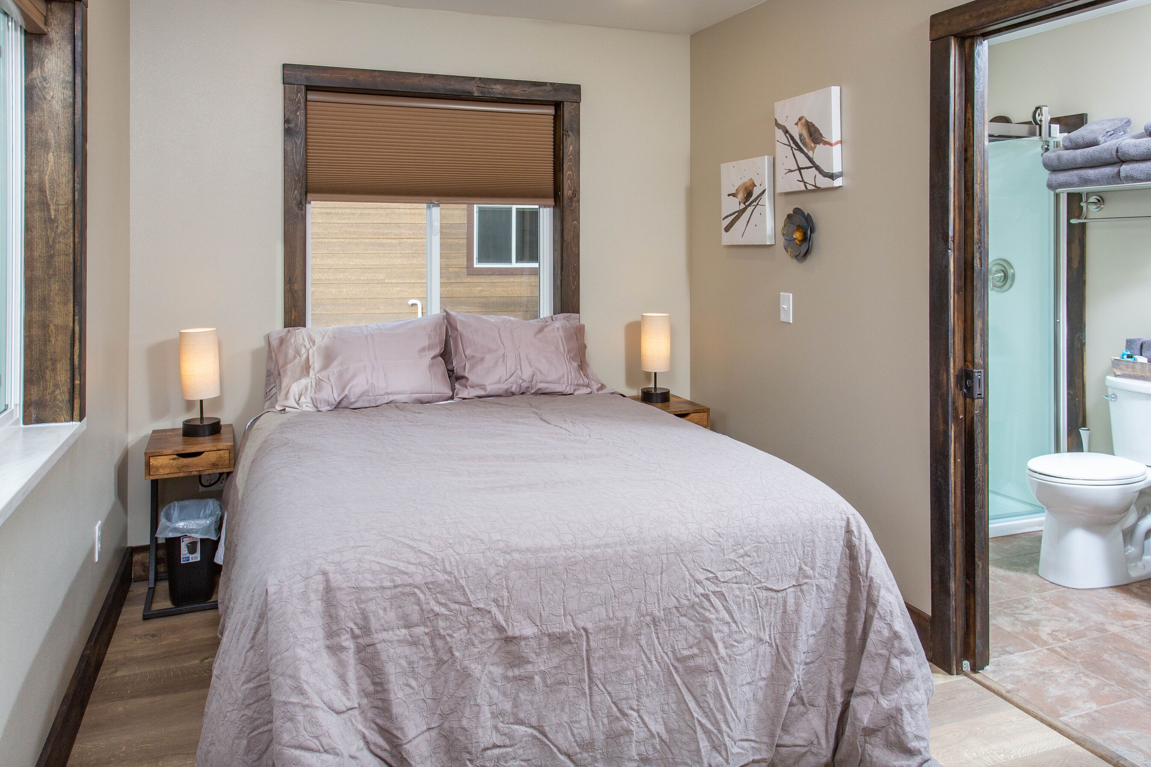 A bedroom with a large bed, two bedside tables with lamps, a window with blinds, and a view of the bathroom.