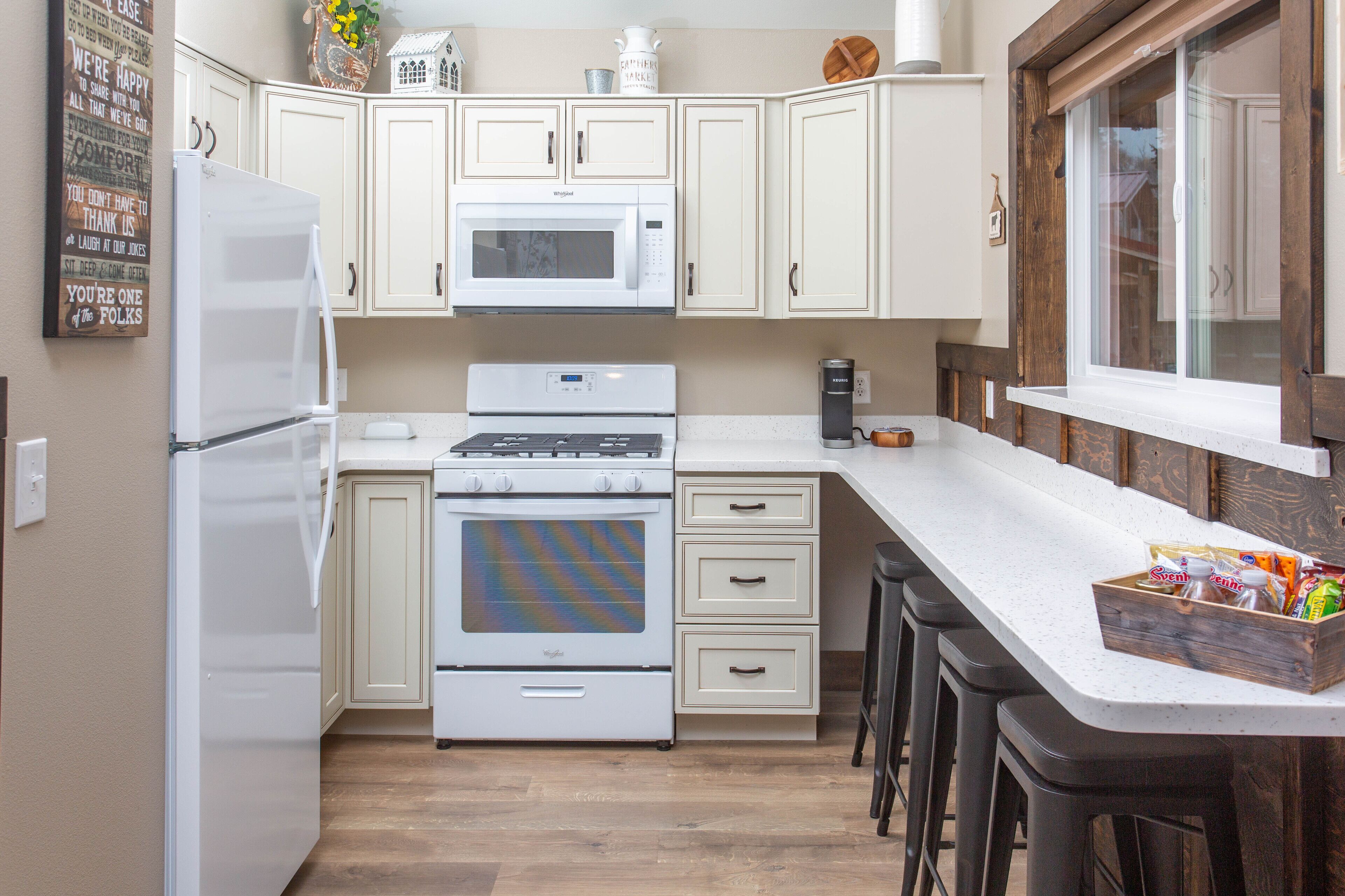 Una cocina con armarios blancos, un microondas, una estufa y un refrigerador. Hay taburetes de bar y una ventana con vistas.