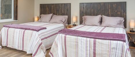 A double room with two beds, each with a wooden headboard, a window, and a nightstand with a lamp.