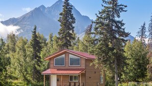 A two-story wooden cabin with a red roof, a porch with seating, and a view of a mountain range. - Alaska Back Country Cottages (Palmer)